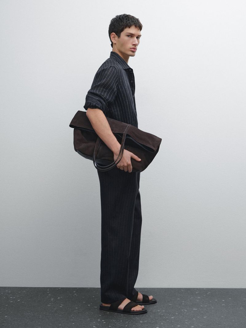 A slender adult man with dark hair is standing in front of a white wall holding a large brown leather bag wearing stylish black sandals.