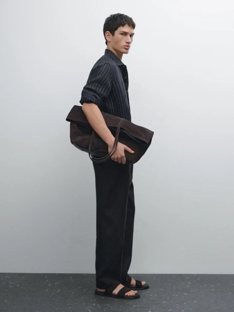 A slender adult man with dark hair is standing in front of a white wall holding a large brown leather bag wearing stylish black sandals.
