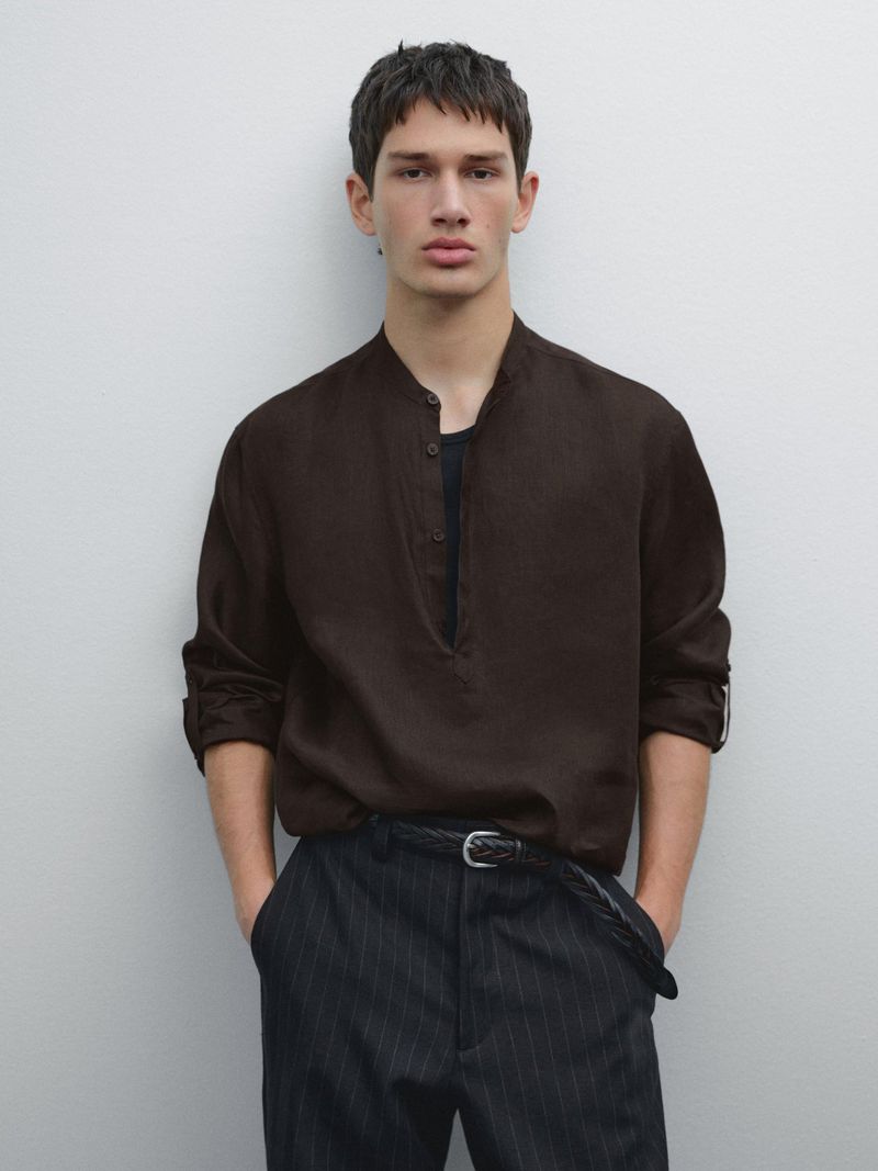 A handsome man poses in a brown button-down shirt and dark pants, standing against a light grey backdrop, showcasing fashion and style.