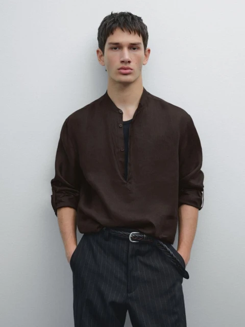 A handsome man poses in a brown button-down shirt and dark pants, standing against a light grey backdrop, showcasing fashion and style.