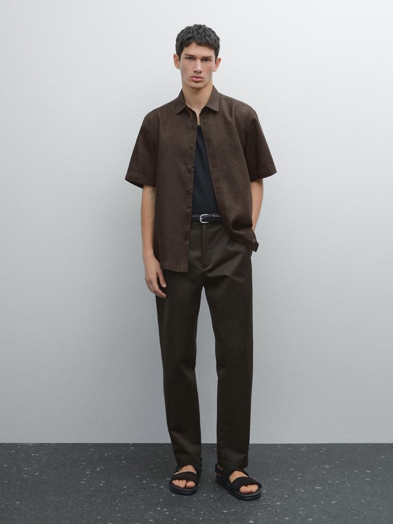 Full shot of a young adult man posing indoors. He wears a short-sleeved brown shirt, brown pants, and black sandals against a neutral background.