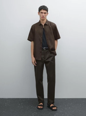 Full-length portrait of a male model wearing a short-sleeve brown shirt, dark trousers, and sandals against a plain studio background.