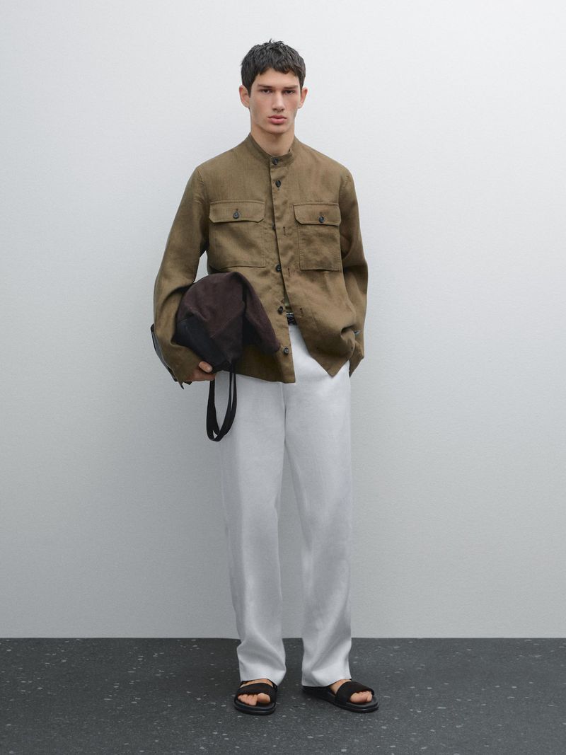 Man in olive green shirt and white pants with black sandals, carrying a brown bag against a plain white background.