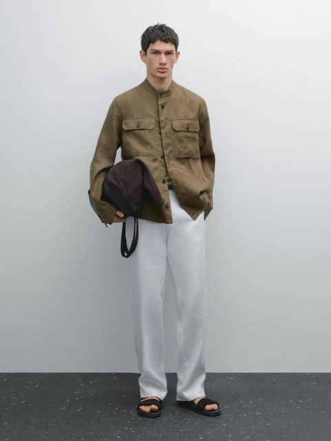 Man in olive green shirt and white pants with black sandals, carrying a brown bag against a plain white background.