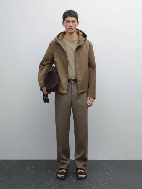 Man in a brown hooded jacket, tan t-shirt, and brown pants, stands in a photo studio holding a dark brown bag.