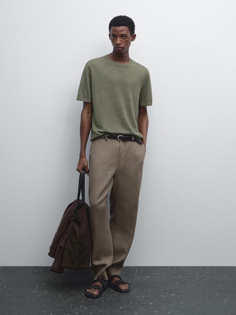 A man in a green short-sleeved t-shirt and tan pants stands against a white wall with a tote bag and brown jacket.