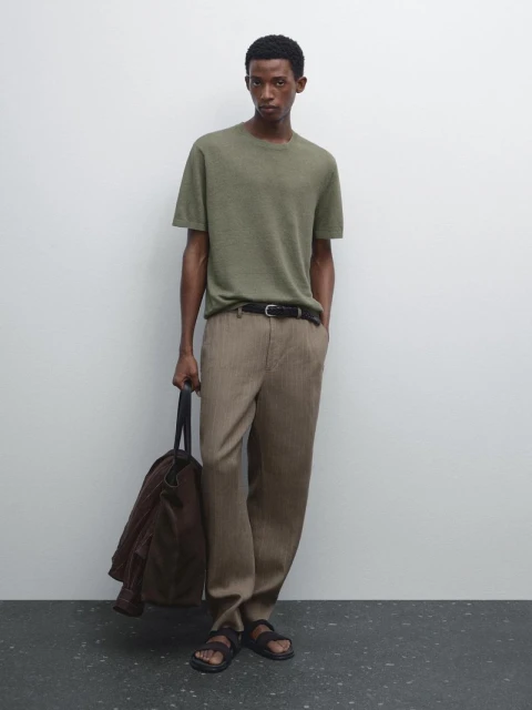 A man in a green short-sleeved t-shirt and tan pants stands against a white wall with a tote bag and brown jacket.