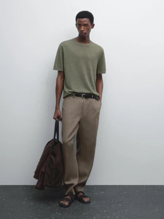 A man in a green short-sleeved t-shirt and tan pants stands against a white wall with a tote bag and brown jacket.
