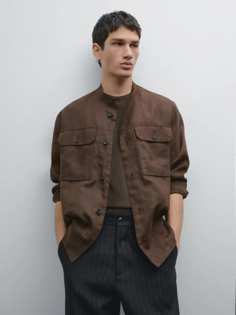A young adult male poses in a brown button-down jacket with patch pockets, a brown t-shirt, and pinstripe pants against a light background.