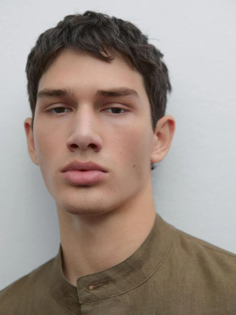 Close-up portrait of a young adult male with dark hair, wearing a tan mandarin-collar shirt against a plain white background.