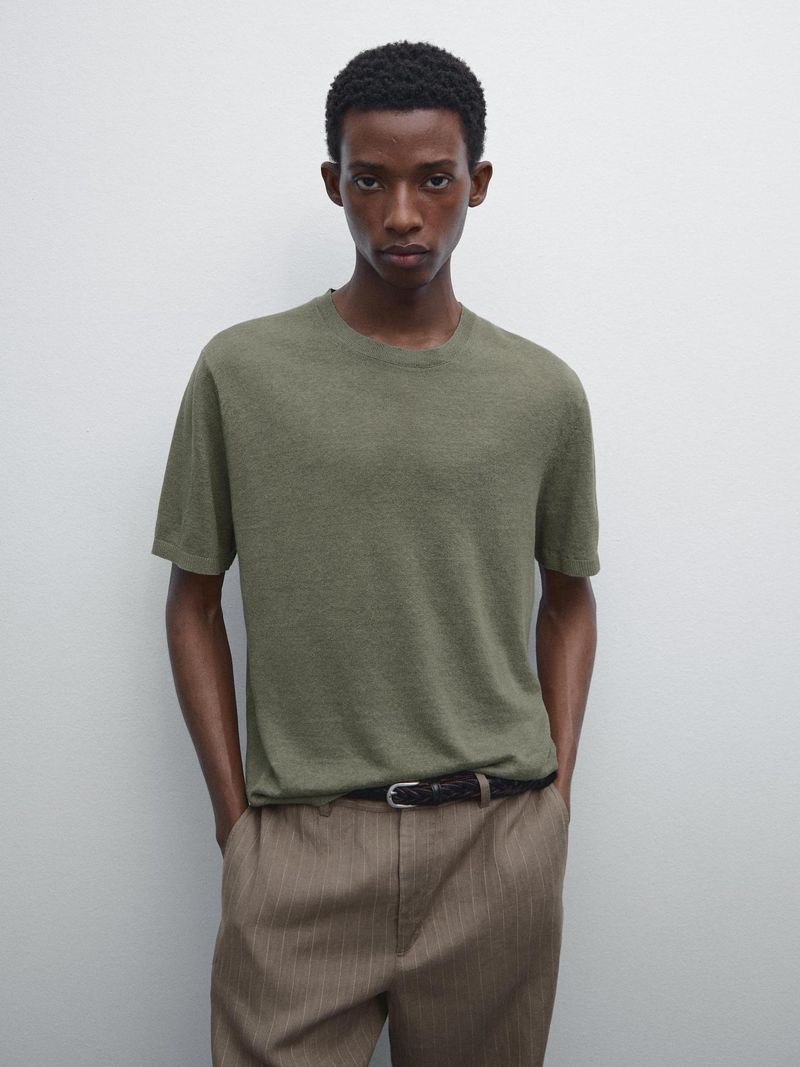 A tan-skinned male model is posing indoors wearing a textured olive-green T-shirt, striped beige trousers, and a black belt in a studio shot.