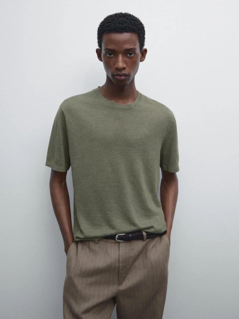 A tan-skinned male model is posing indoors wearing a textured olive-green T-shirt, striped beige trousers, and a black belt in a studio shot.