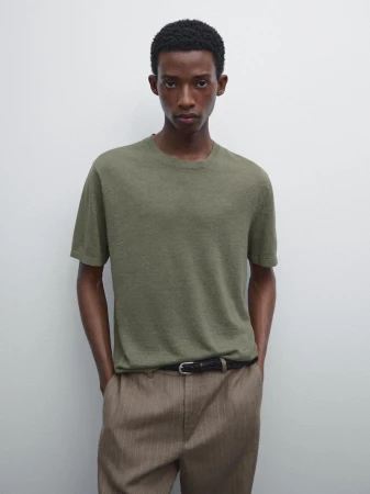 A tan-skinned male model is posing indoors wearing a textured olive-green T-shirt, striped beige trousers, and a black belt in a studio shot.