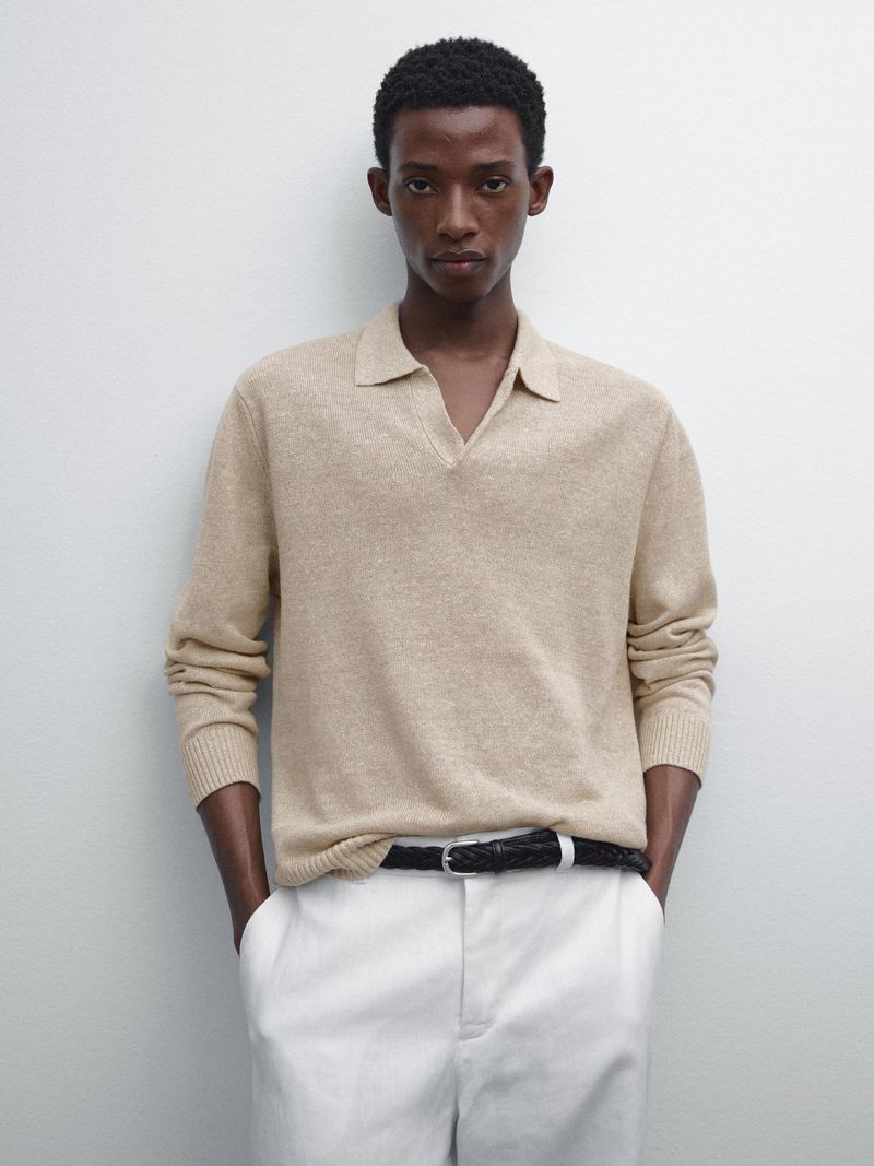 Studio shot of a Black man in a beige polo sweater, white pants, and black belt, posing against a neutral backdrop for a fashion editorial.
