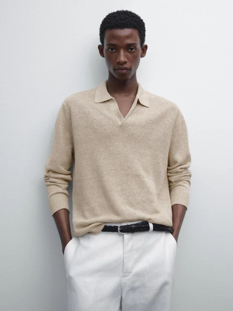 Studio shot of a Black man in a beige polo sweater, white pants, and black belt, posing against a neutral backdrop for a fashion editorial.