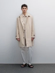 Full-length portrait of a male model wearing a light beige coat and trousers against a neutral grey studio wall.