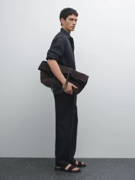 Male model in dark pinstripe shirt and trousers carrying a large brown suede bag against a seamless light background