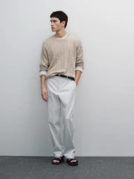 Model standing against a plain light wall wearing a beige cable-knit sweater and light trousers