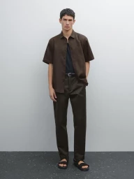Full-length portrait of a male model wearing a short-sleeve brown shirt, dark trousers, and sandals against a plain studio background.