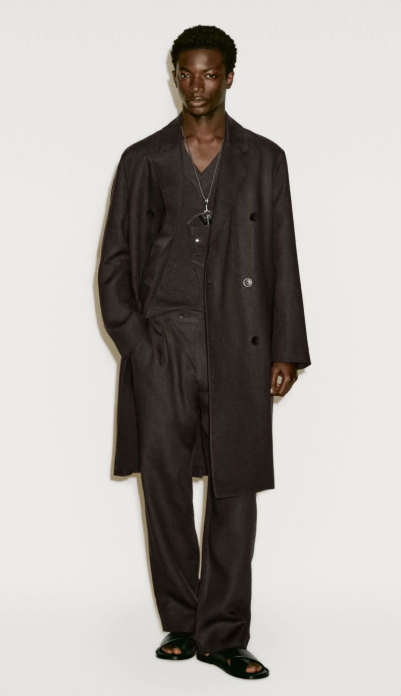 Dark-skinned male fashion model poses in a long, dark overcoat, matching pants, and sandals against a neutral backdrop in a studio shot.