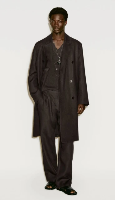 Dark-skinned male fashion model poses in a long, dark overcoat, matching pants, and sandals against a neutral backdrop in a studio shot.