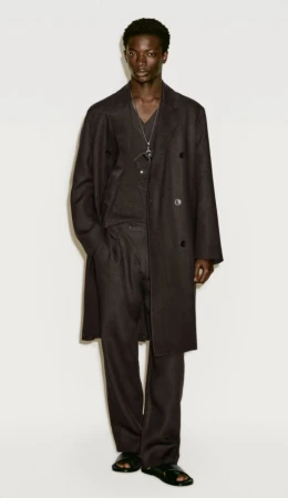 Dark-skinned male fashion model poses in a long, dark overcoat, matching pants, and sandals against a neutral backdrop in a studio shot.