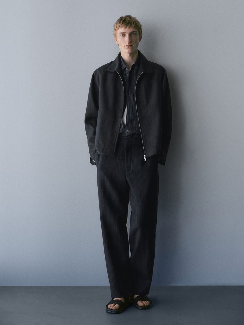 Full-length portrait of a male model wearing a dark jacket, black trousers, and sandals against a grey wall