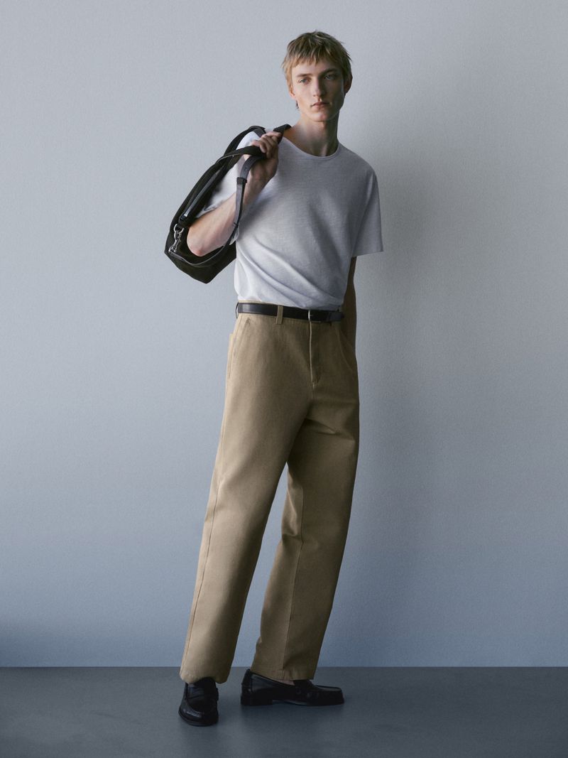 Full-length shot of a young adult man in a white t-shirt, khaki pants, and black loafers, carrying a black leather bag against a grey backdrop.