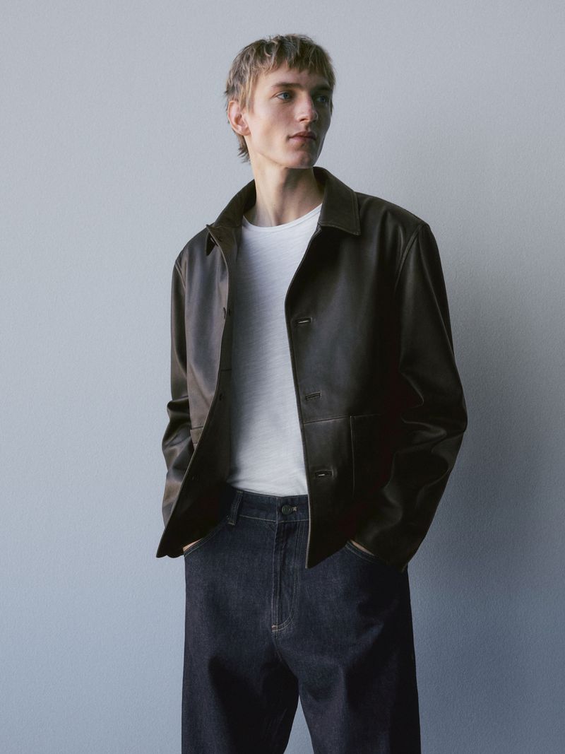 A young male model wearing a dark leather jacket, white t-shirt, and dark denim jeans against a seamless grey backdrop.