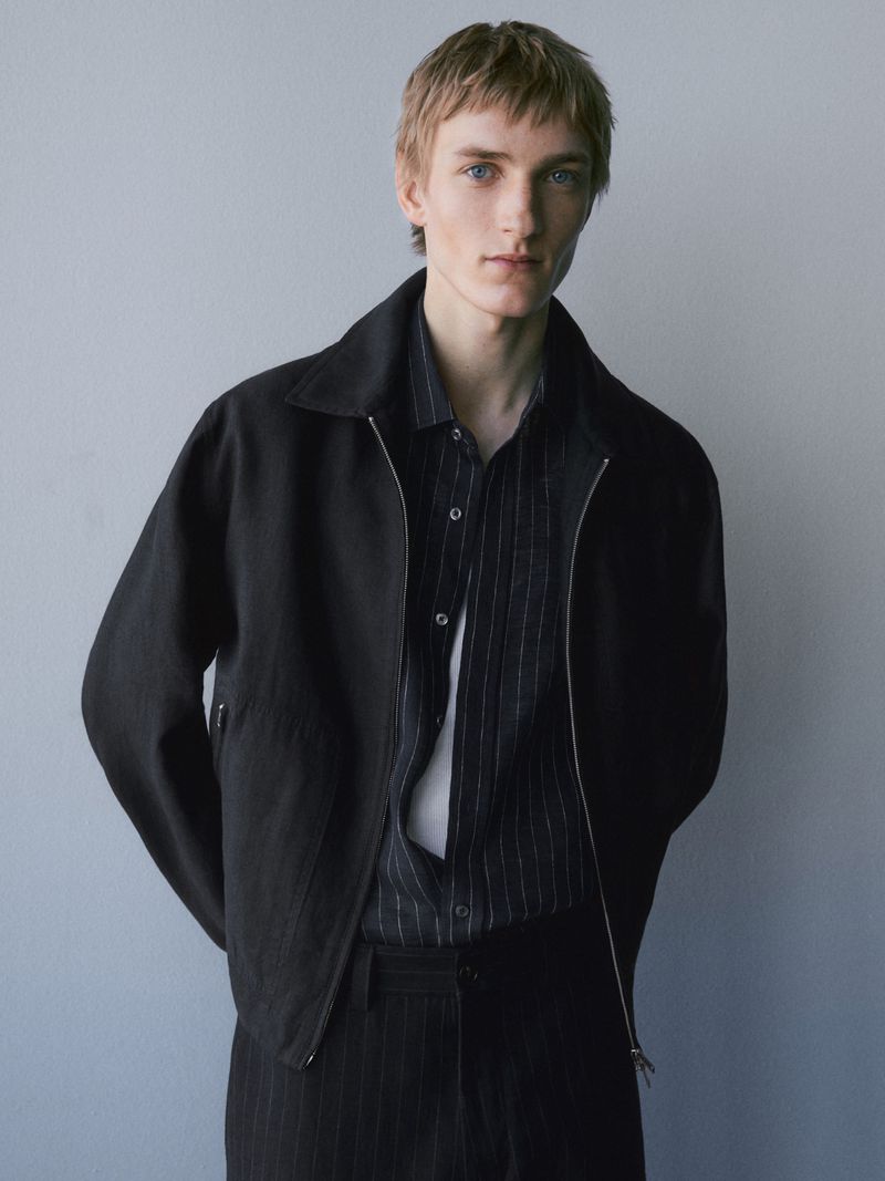 A young male model wearing a black jacket and striped shirt against a neutral grey seamless background.