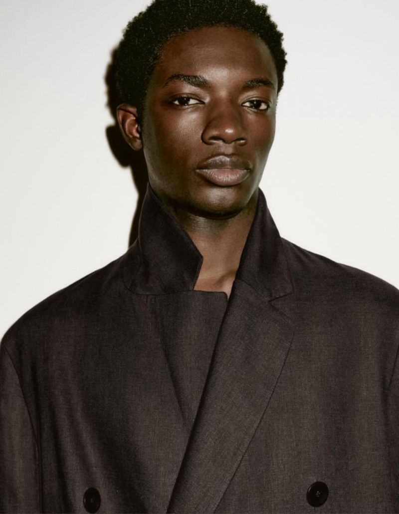 Portrait of an adult male model with dark skin wearing a dark-colored double-breasted jacket against a neutral studio backdrop.