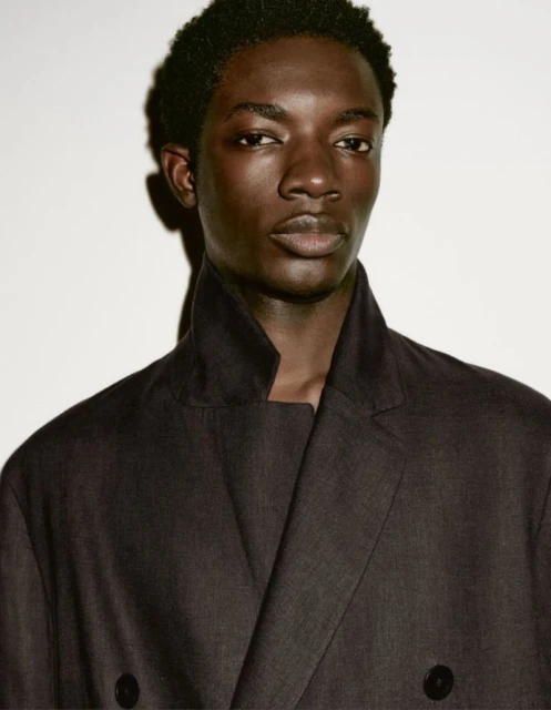 Portrait of an adult male model with dark skin wearing a dark-colored double-breasted jacket against a neutral studio backdrop.