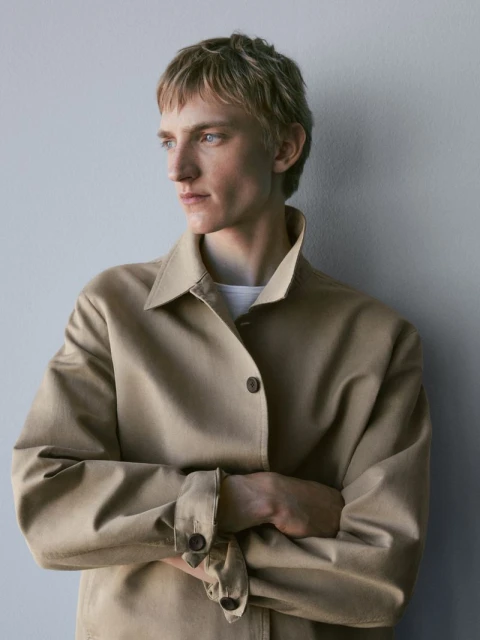 Portrait of a blond male model in a tan jacket with arms crossed, looking to the side against a neutral gray studio backdrop.