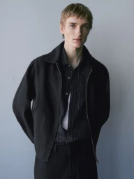 A young male model wearing a black jacket and striped shirt against a neutral grey seamless background.