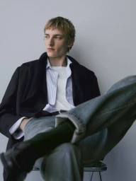 Young male model seated with legs crossed wearing a striped shirt, dark jacket, and blue jeans against a plain grey background