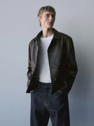 A young male model wearing a dark leather jacket, white t-shirt, and dark denim jeans against a seamless grey backdrop.
