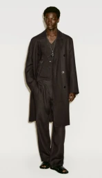 Black man standing against a white backdrop wearing a dark wool coat, trousers, and dark sandals.