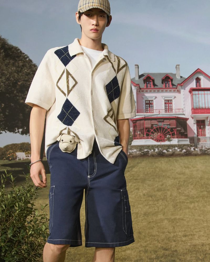 Young man in a patterned shirt and shorts stands outdoors in front of a house. He wears a hat and a dog-shaped coin purse.