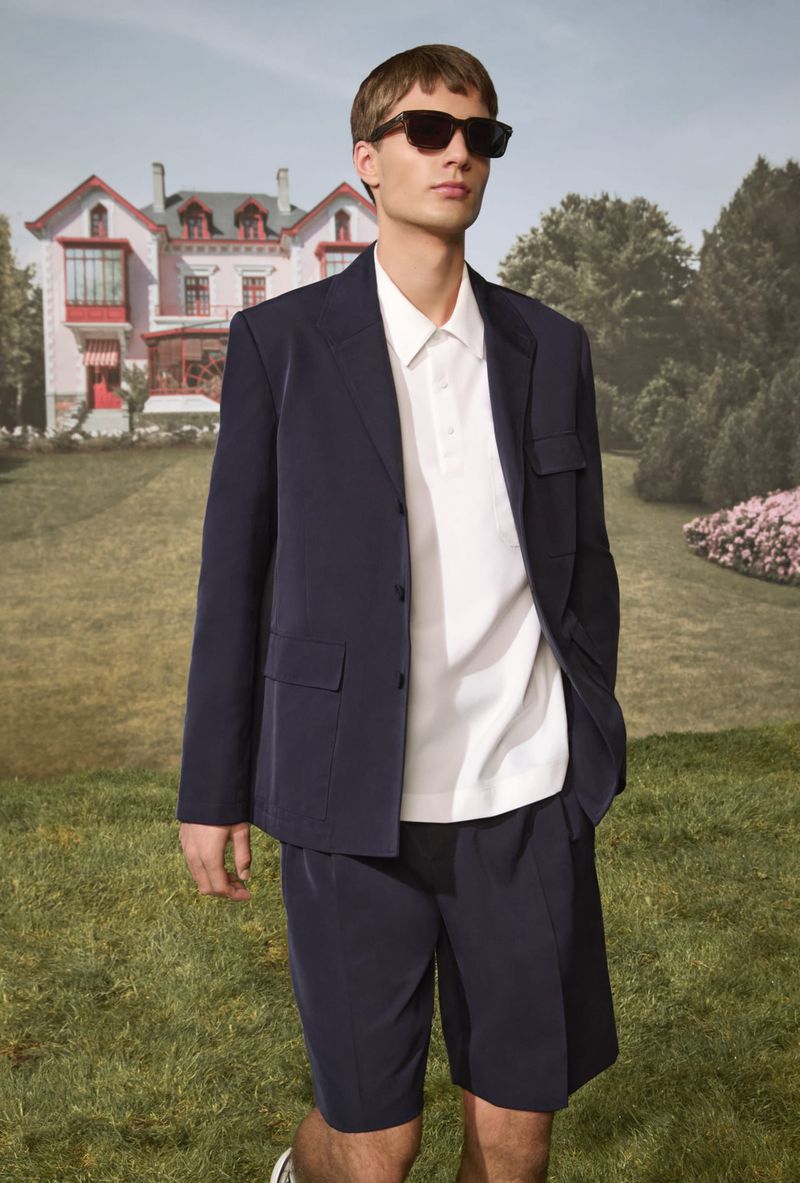 Man in a navy blazer and shorts, wearing sunglasses, stands in a grassy field with a large house in the background on a sunny day.