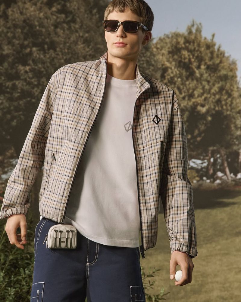 A young man in a plaid jacket and blue shorts stands outside on a sunny day, holding a golf ball, with trees in the background.
