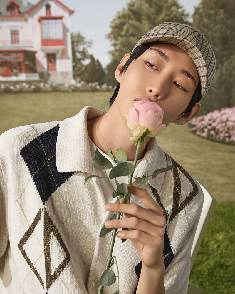 Close-up of an adult male model in a hat and sweater, holding a rose in front of a backdrop with a pink house.