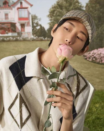 Close-up of an adult male model in a hat and sweater, holding a rose in front of a backdrop with a pink house.