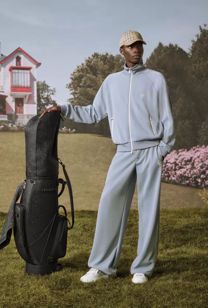 An adult male model poses in a light blue tracksuit with a golf bag on a green lawn, with a pink and white house in the background.