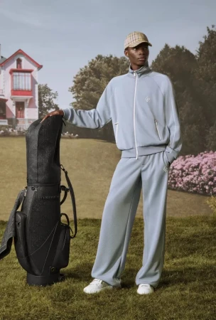 An adult male model poses in a light blue tracksuit with a golf bag on a green lawn, with a pink and white house in the background.