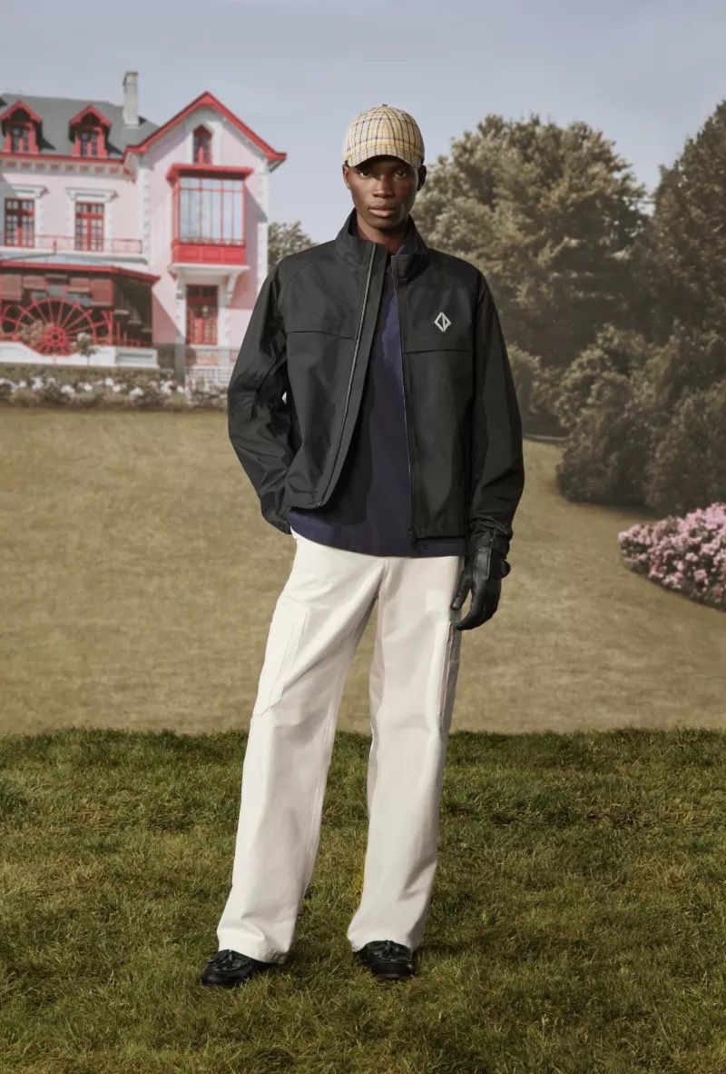 Man posing outdoors wearing black jacket, cream pants, and plaid cap, with a historic building and trees in the background.