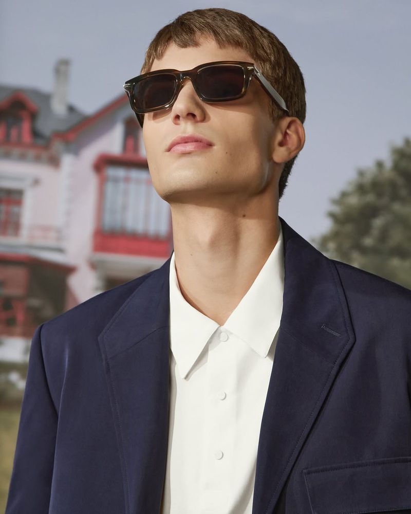 Close-up portrait of a male model wearing dark sunglasses, a white collared shirt, and a navy blue blazer.