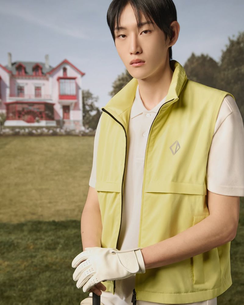Asian man poses in a lime green vest and golf glove outdoors with a golf club and a pink house in the background on a grassy lawn.