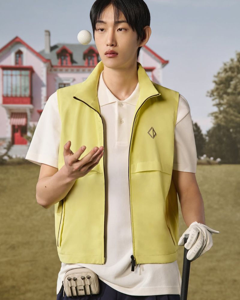Male model in yellow vest poses with golf ball and club against pink house and blue sky on green golf course backdrop.