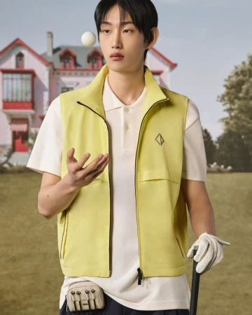 Male model in yellow vest poses with golf ball and club against pink house and blue sky on green golf course backdrop.
