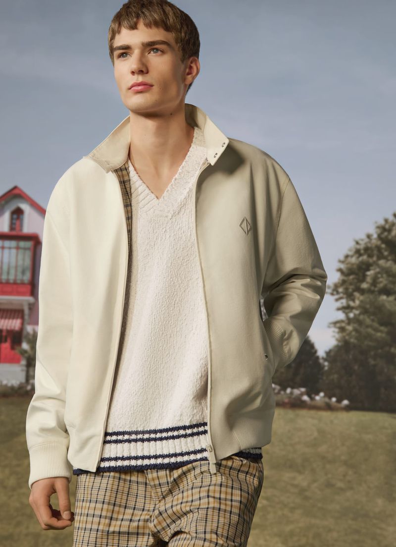 Young man poses wearing a cream-colored jacket, sweater, and plaid pants, outdoors in a field with a house in the background.